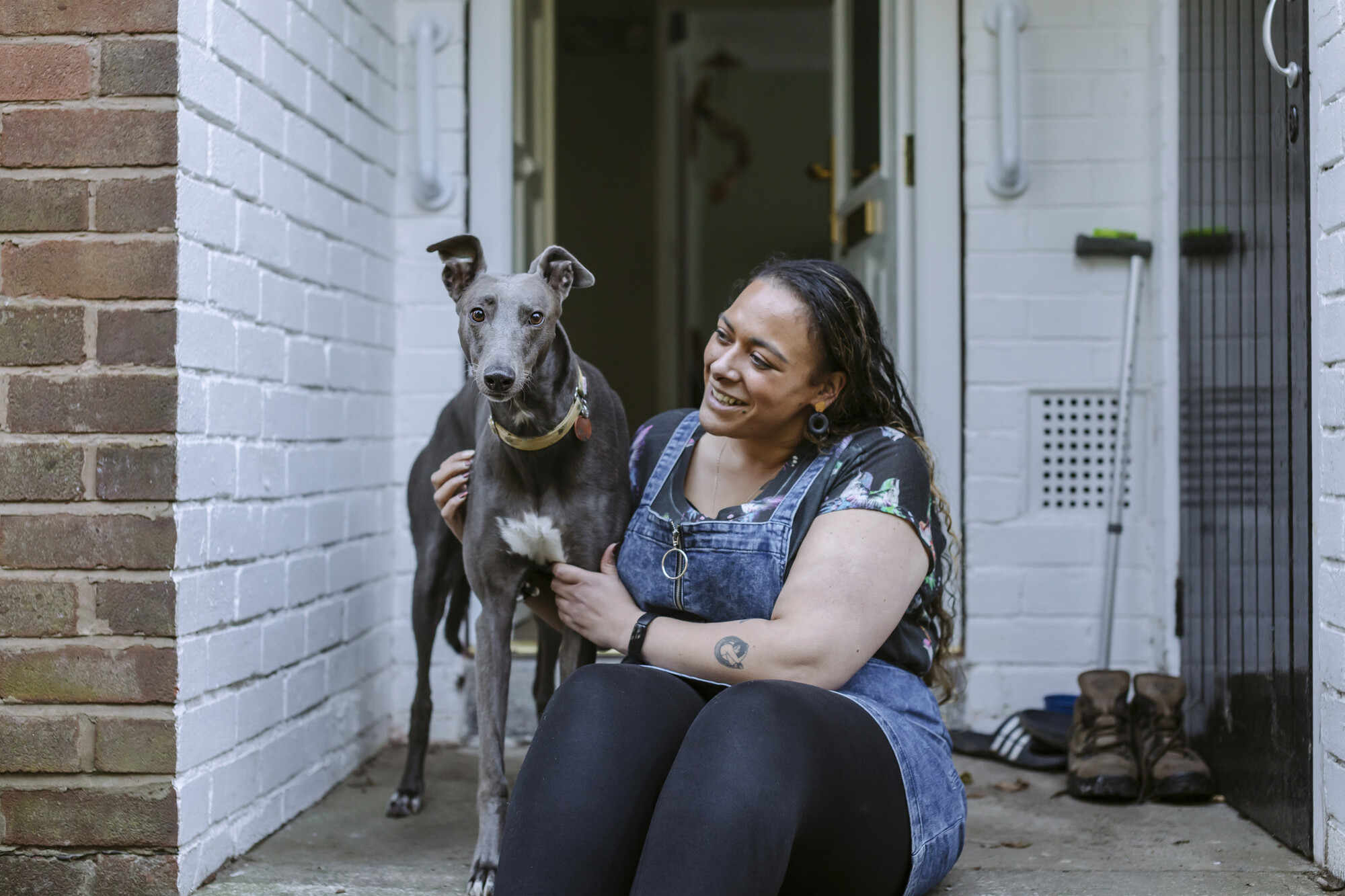 Greyhound Bleu standing on her doorstep with owner Amy sitting beside her, smiling