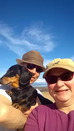 A man and woman hold a black and tan dachshund with views of a sandy beach and blue sky behind them