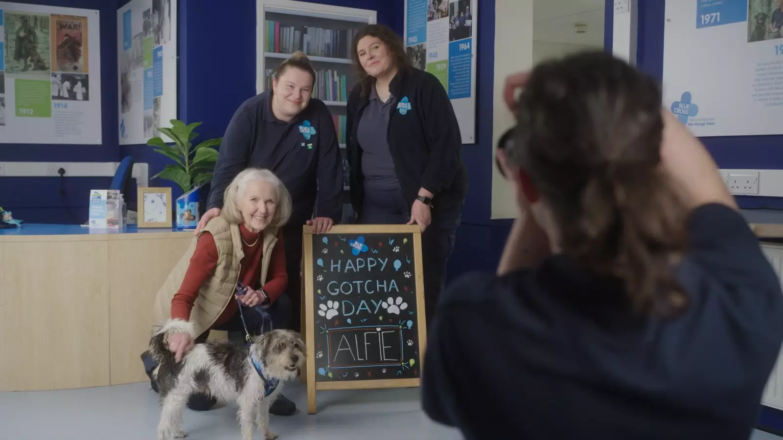 A new owner with dog, Alfie, besides a sign reading 'Happy Gotcha Day Alfie' with some Blue Cross staff